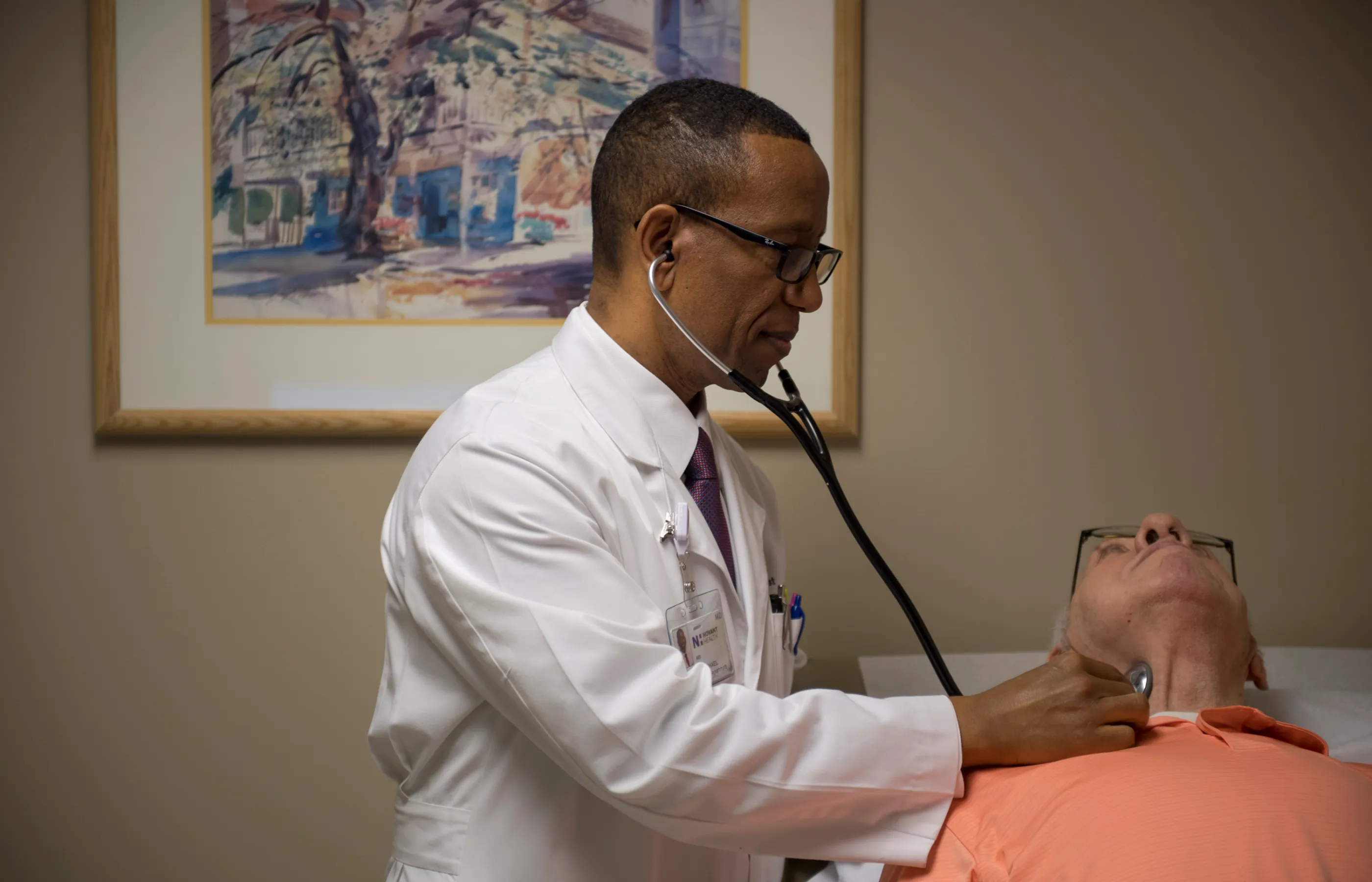 Dr. Michael is in a exam room with a patient. The patient is lying down on the exam table as Dr. Michael places his stethoscope on their neck to listen. 