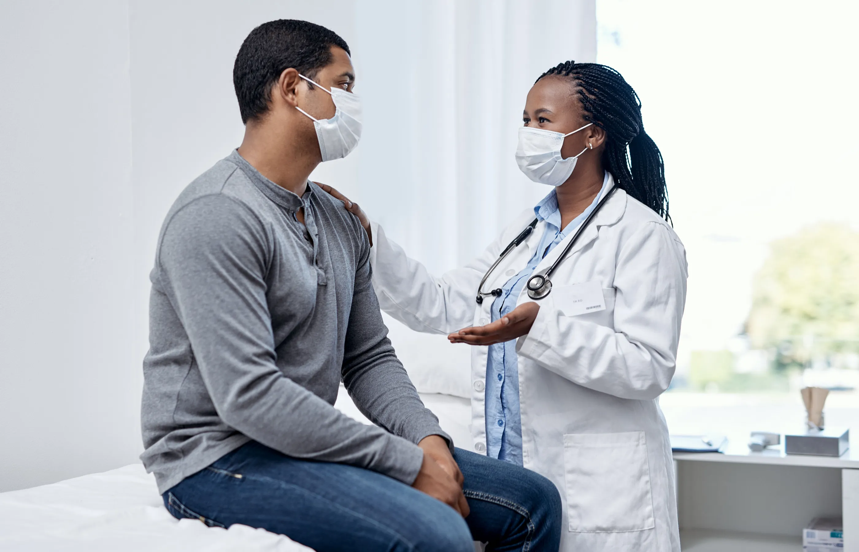 A doctor and patient are both wearing masks during their visit. The patient is sitting on the exam table as the provider is talking and resting her hand on his shoulder. 