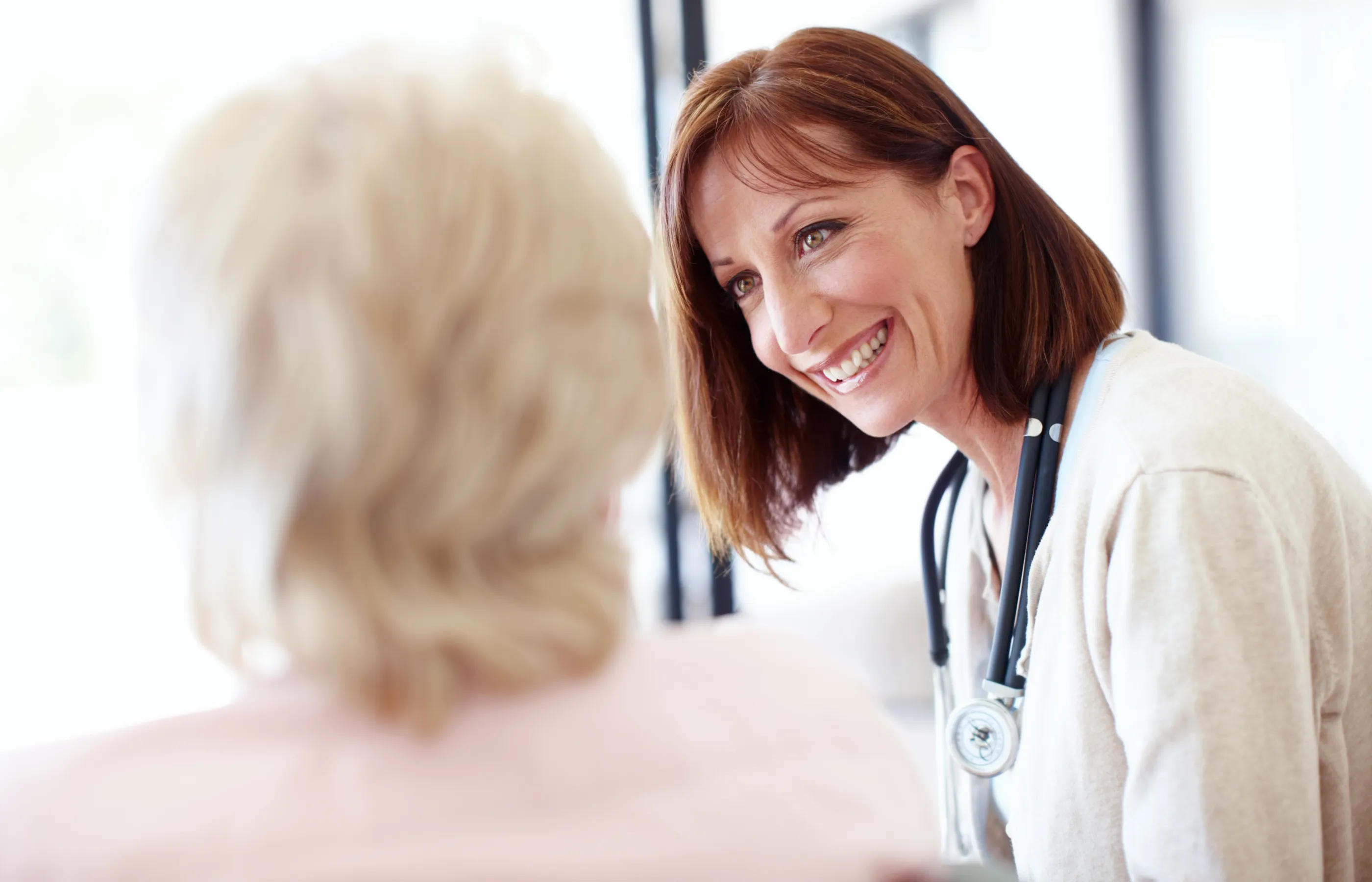 A doctor is sitting with a lab coat and stethoscope around her neck. She is smiling and looking at her patient. 