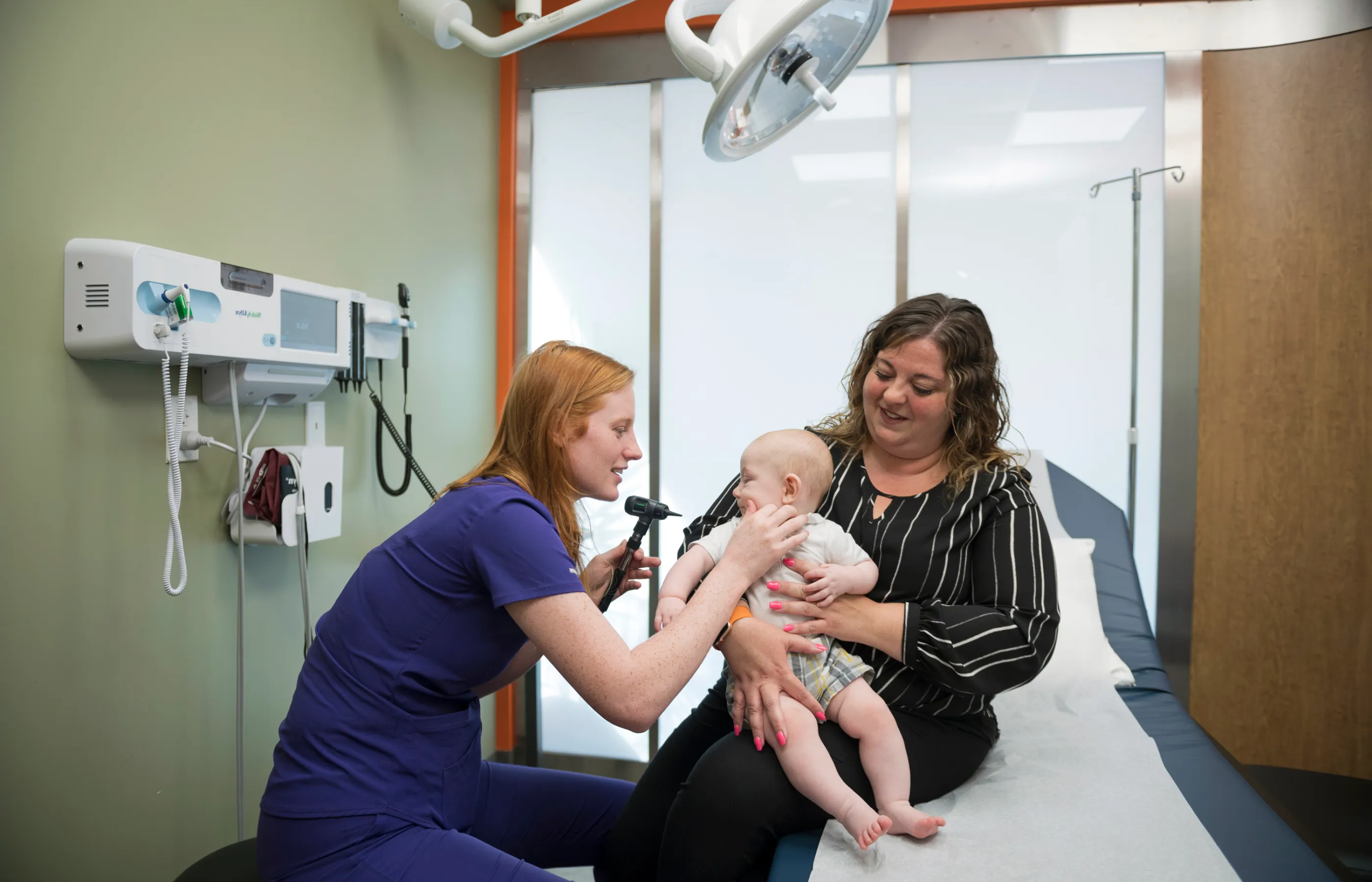 A nurse checking a baby's ear while the baby is held by it's mother.