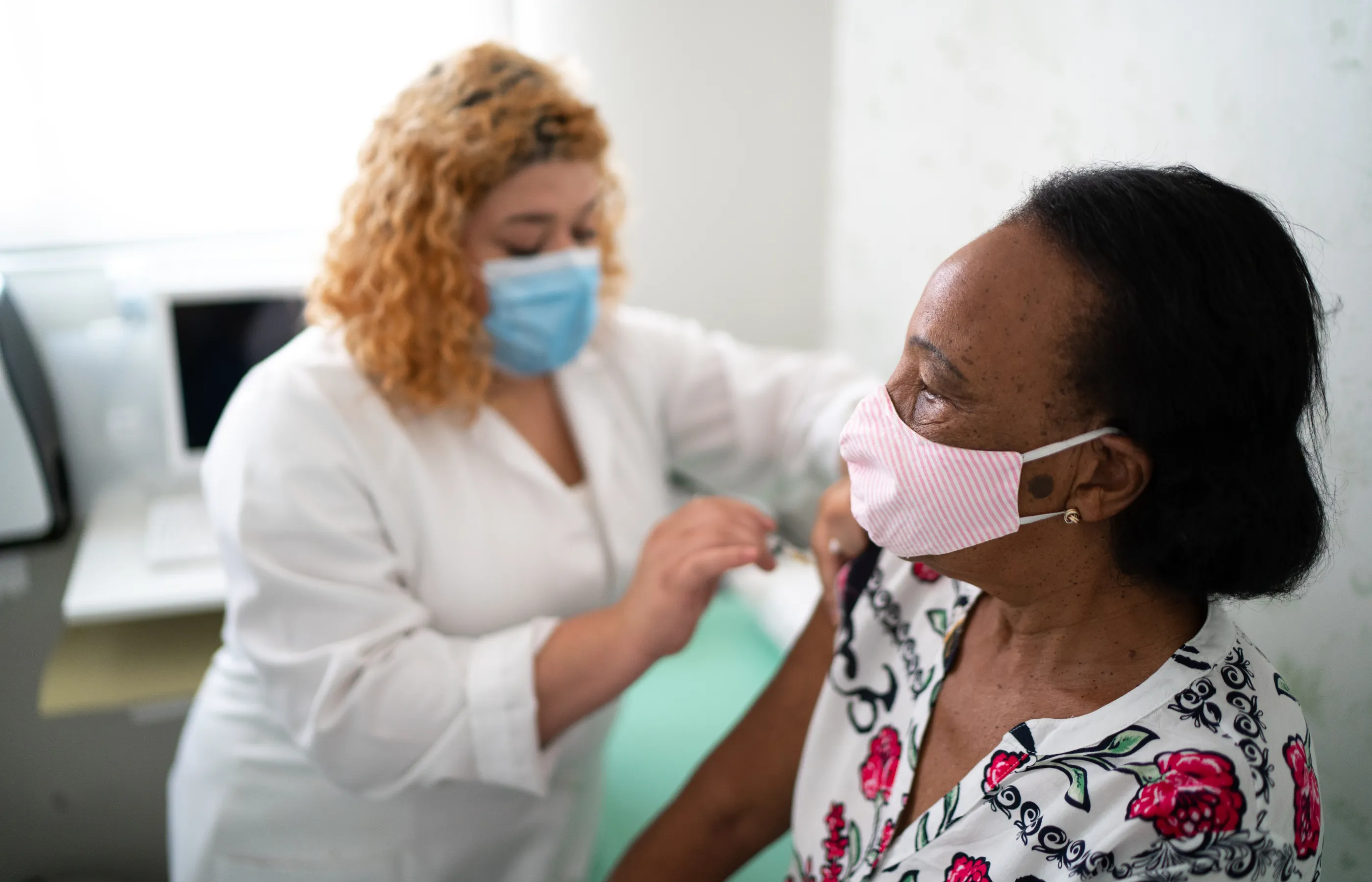 A woman is sitting on an exam table as she recieves a vaccine from her health care provider. Both the patient and provider are wearing masks. 