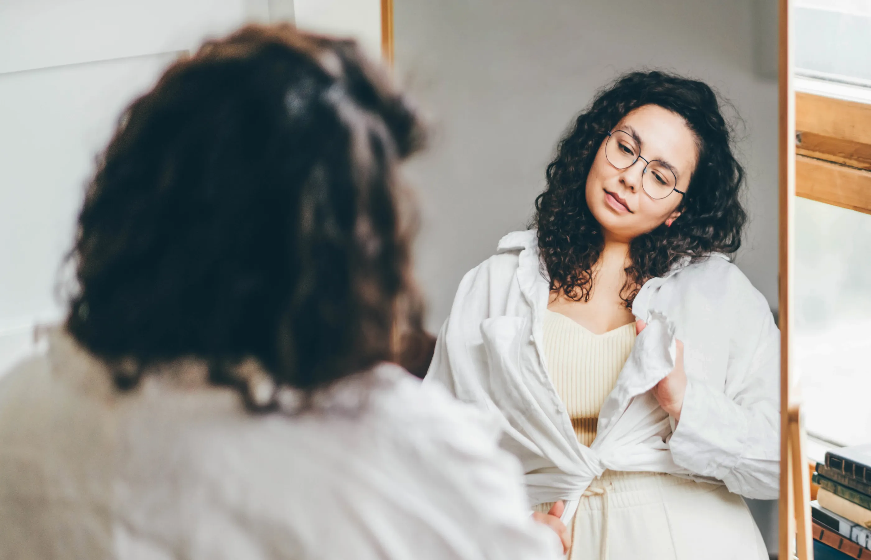 A young woman is looking at herself in the mirror as she gets dressed.