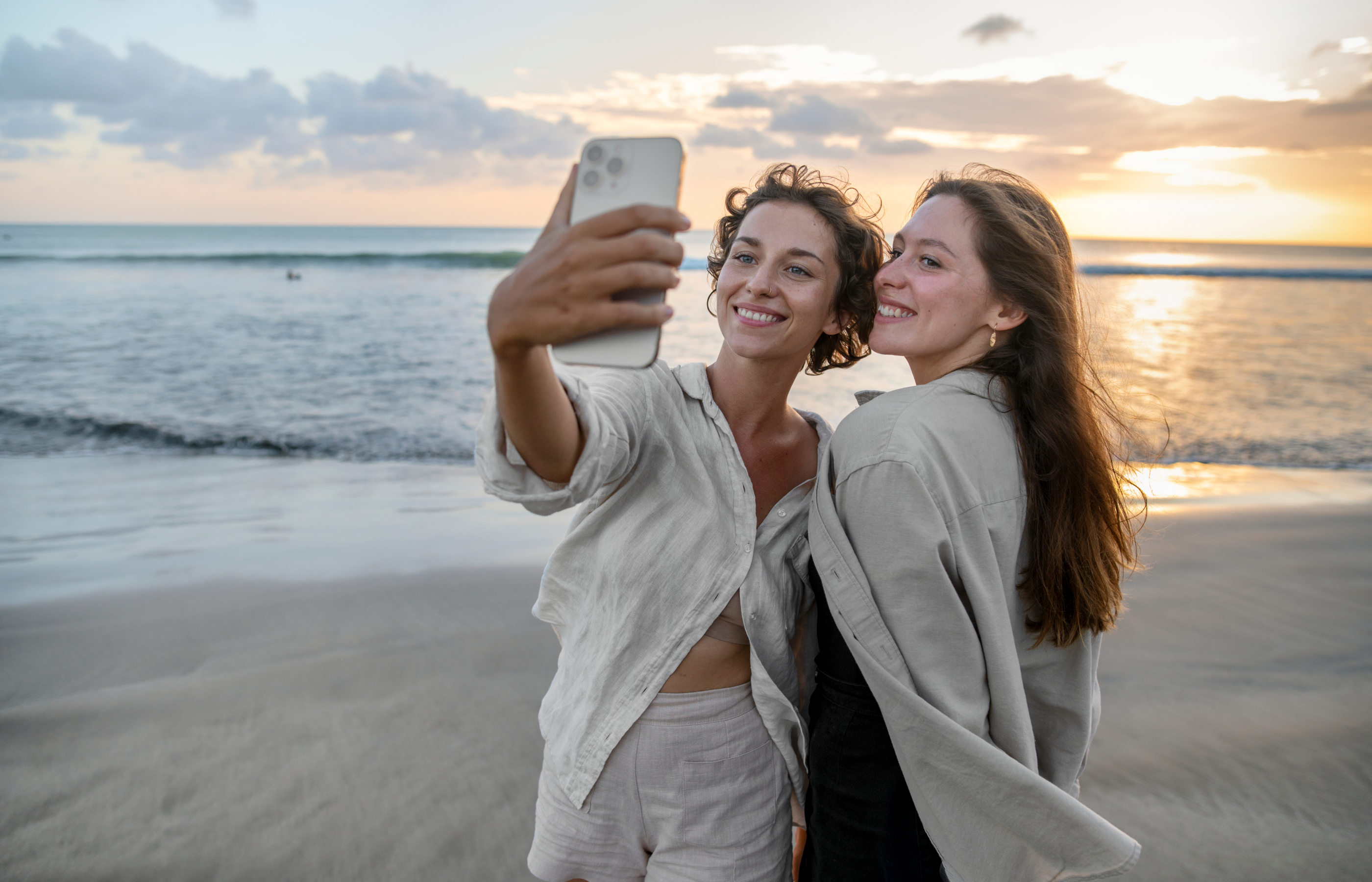 Two women taking a selfie using a smart phone with a sunrise behind them at the beach.