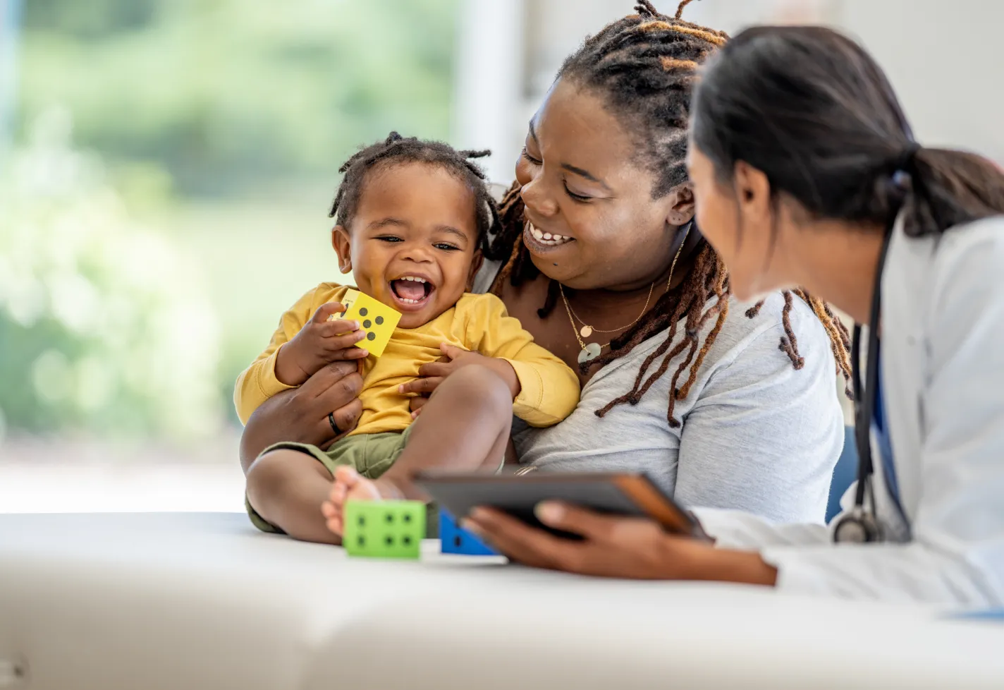 A mom, her toddler, and a provider holding a tablet smile as the baby plays with dice blocks