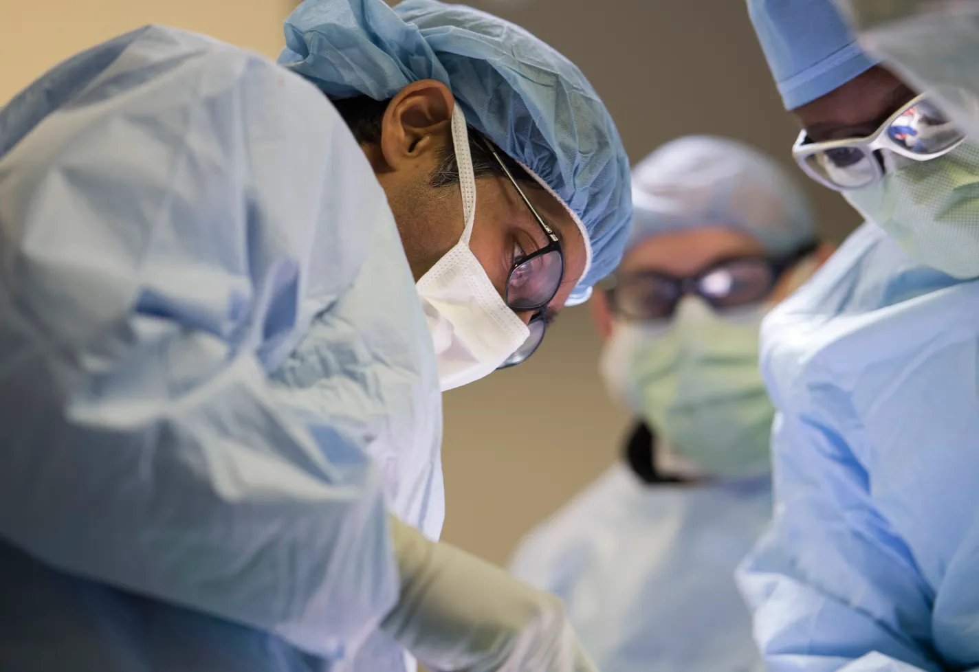 3 surgeons in operating masks, glasses, and caps look down at a procedure 3 surgeons in operating masks, glasses, and caps look down at a procedure