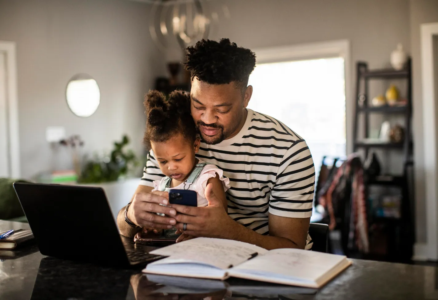 Father is sitting with his daughter at the counter. They are sitting in front of a laptop as he is looking for information on his phone.