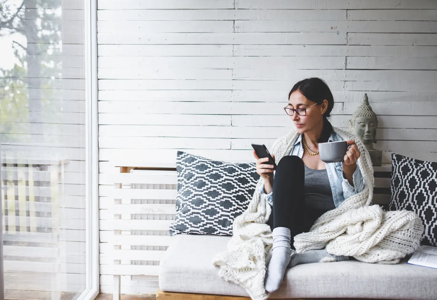A woman is sitting on the couch, wearing a blanket, drinking coffee while on her smartphone. A woman is sitting on the couch, wearing a blanket, drinking coffee while on her smartphone.