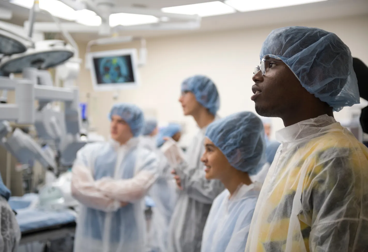 A group of students are wearing personal protective equipment (PPE) as they stand and observe in an operating room. A group of students are wearing personal protective equipment (PPE) as they stand and observe in an operating room.