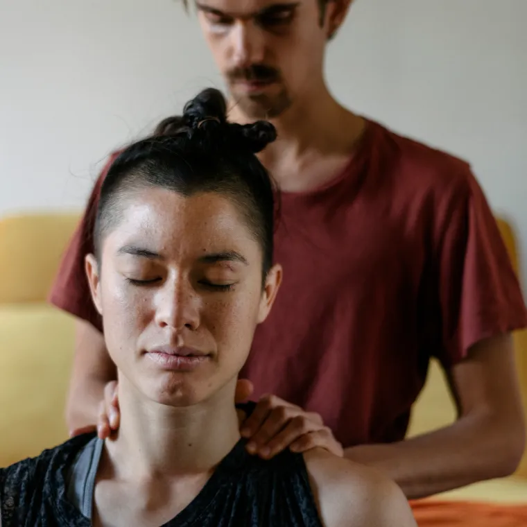 A woman is sitting on the floor with her eyes closed as her partner massages her shoulders. 
