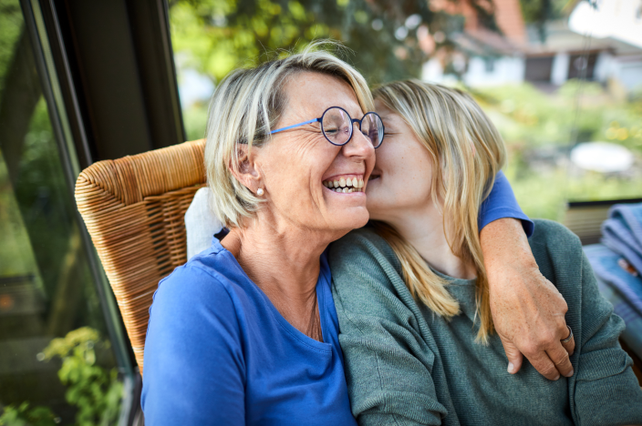 A woman is hugging her adult daughter, as she leans in to kiss her cheek. 