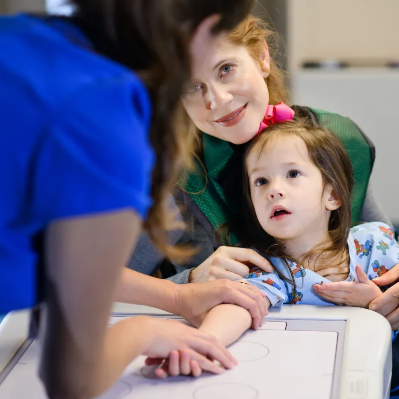 A young girl is sitting on her moms lap while a Novant Health team member positions her arm for an x-ray. A young girl is sitting on her moms lap while a Novant Health team member positions her arm for an x-ray.